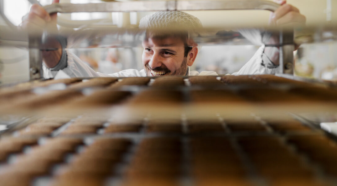 Picture of professional male baker man in white work uniform . Standing in front of the shelves full with fresh baked cookies.